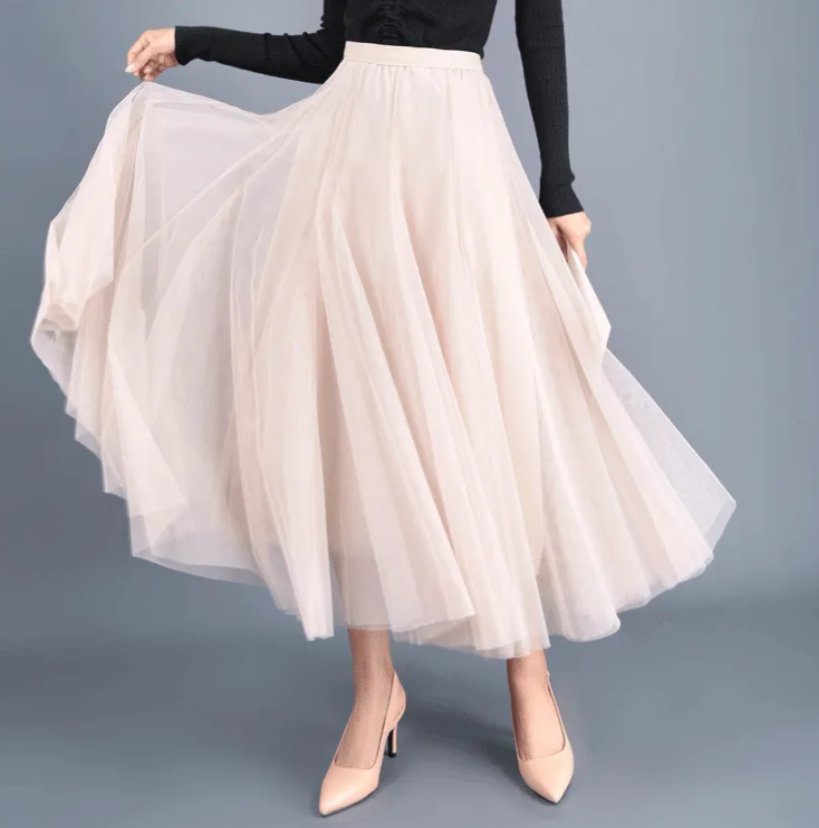 Woman in a flowing beige tulle skirt and black top, twirling to show the fullness of the ankle-length skirt paired with beige high heels.