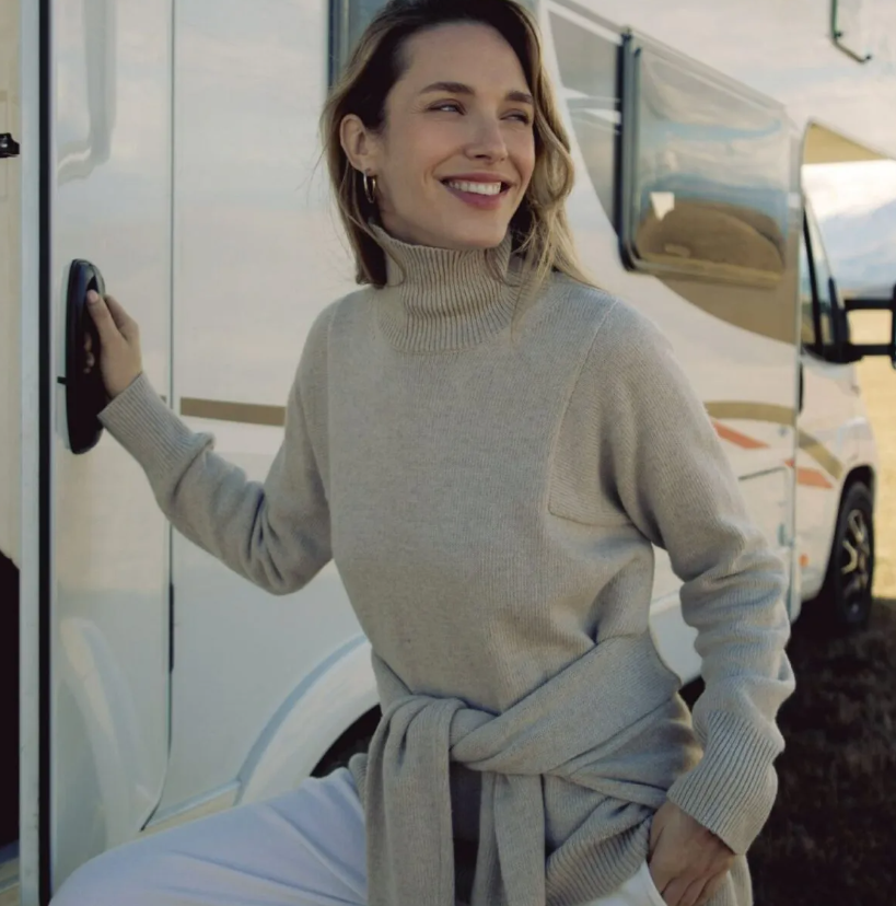 Smiling woman stepping out of an RV, wearing a chic gray turtleneck sweater tied at the waist.